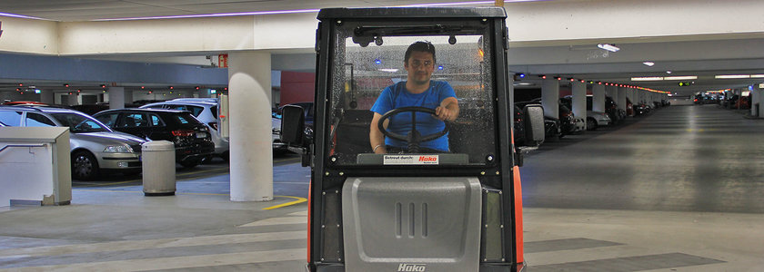 Person in blauem T-Shirt sitzt in einem kleinen Reinigungsfahrzeug in einer Tiefgarage mit mehreren geparkten Autos